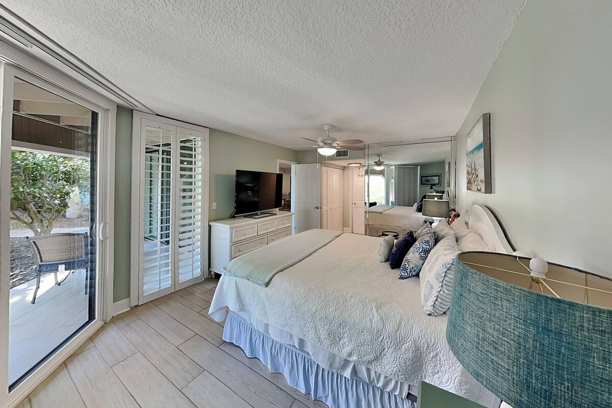 A bedroom with a large bed, a TV, a ceiling fan, and a sliding glass door leading to an outdoor area.