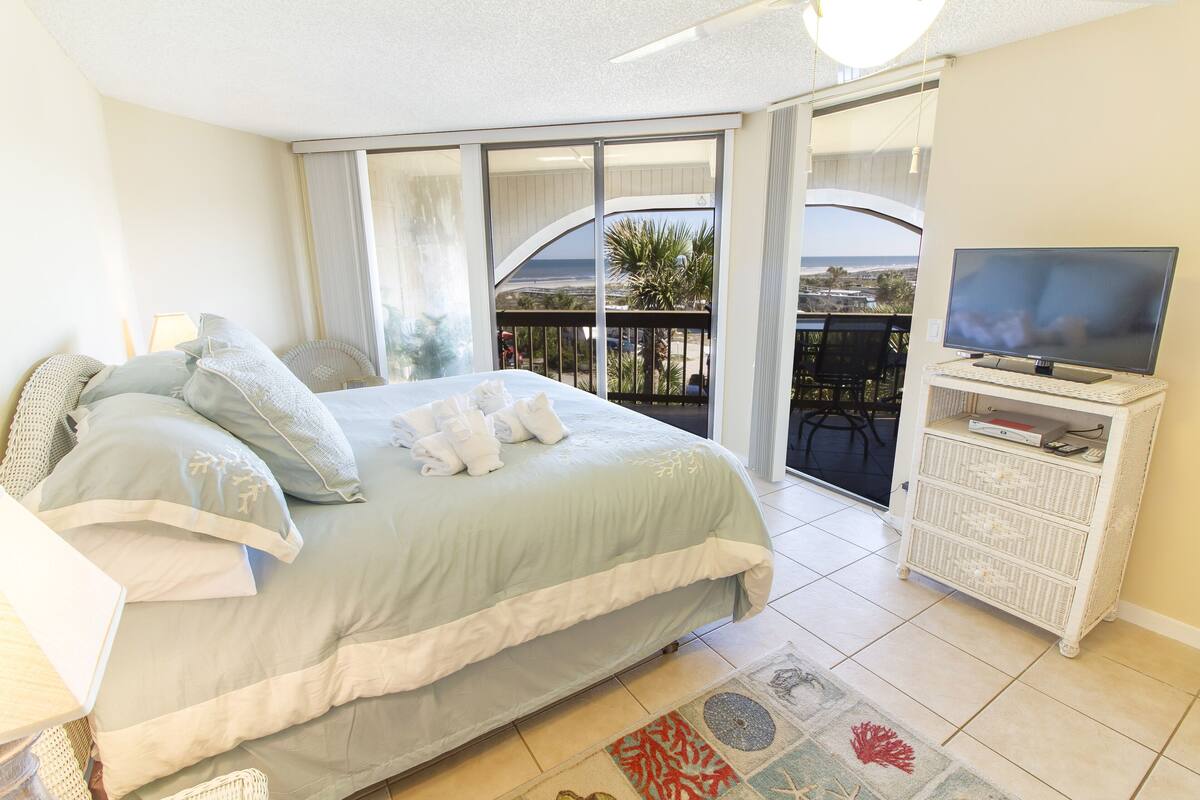 A bedroom with a bed, a television, and a balcony with a view of the beach.