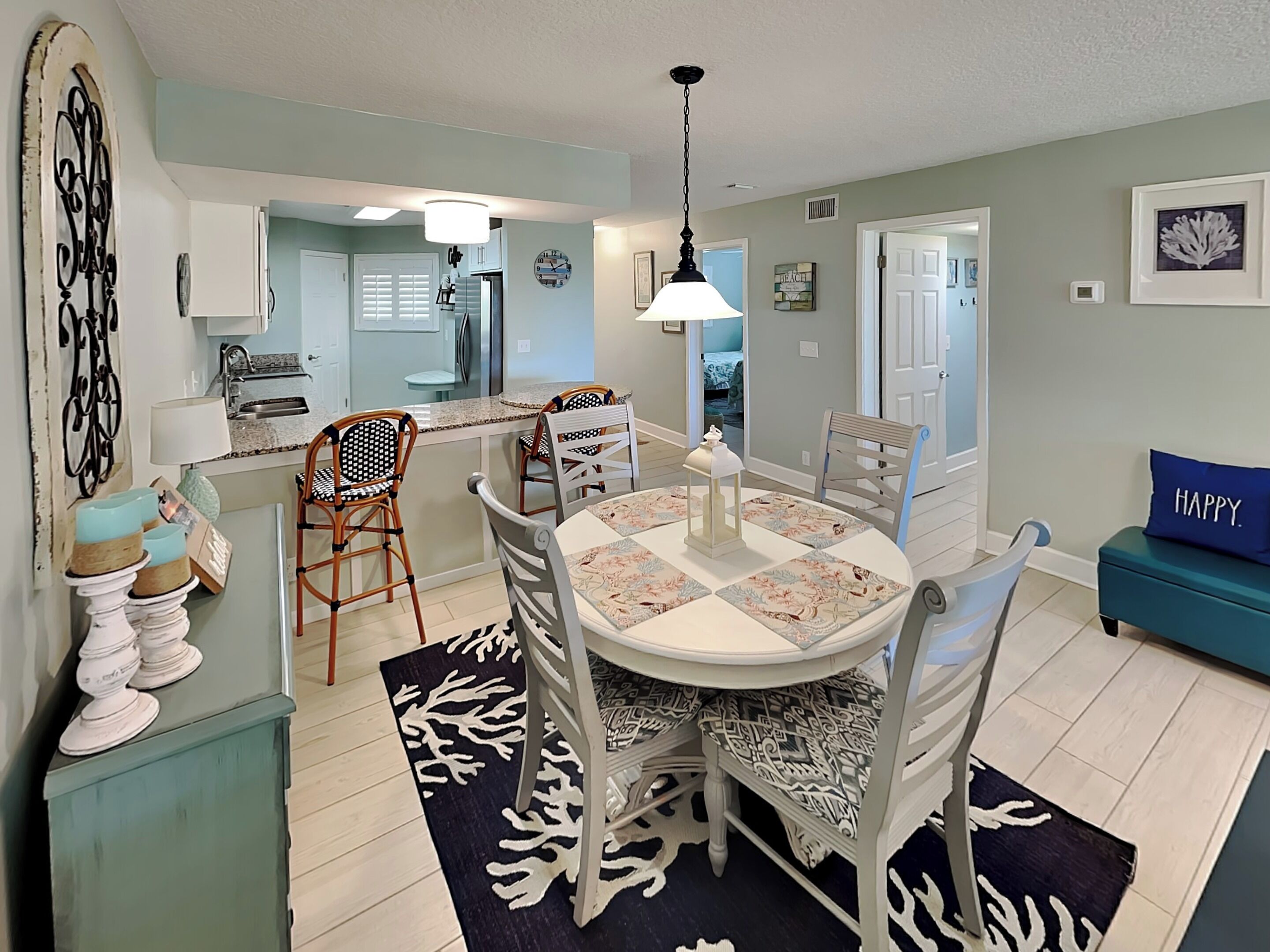 A dining area with a round table, chairs, and a rug. There is a kitchen in the background with cabinets and a sink.