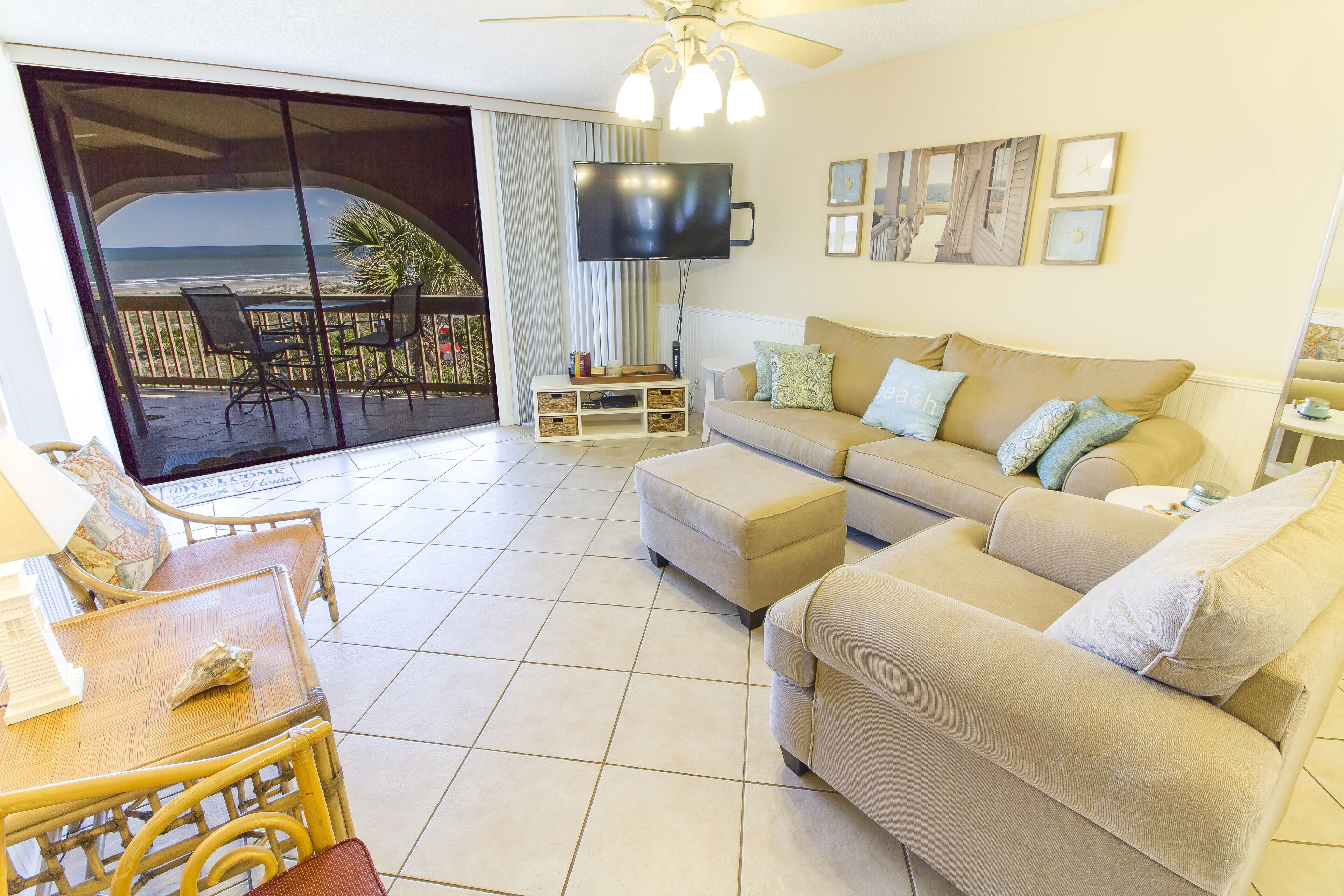 Ein Wohnzimmer mit einer Couch, Hocker und einem Tisch mit Stühlen. Ein Fernseher ist an der Wand montiert und ein Balkon bietet Blick aufs Meer.