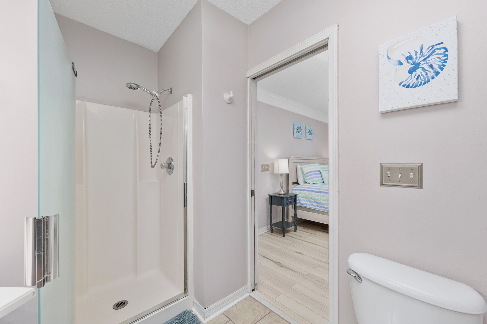 A modern bathroom with a glass shower enclosure, a white toilet, and a bedside table with a lamp.