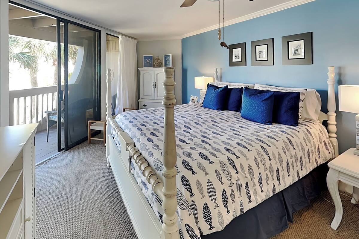A bedroom with a bed, a ceiling fan, a nightstand, a lamp, and a view of the outdoors.