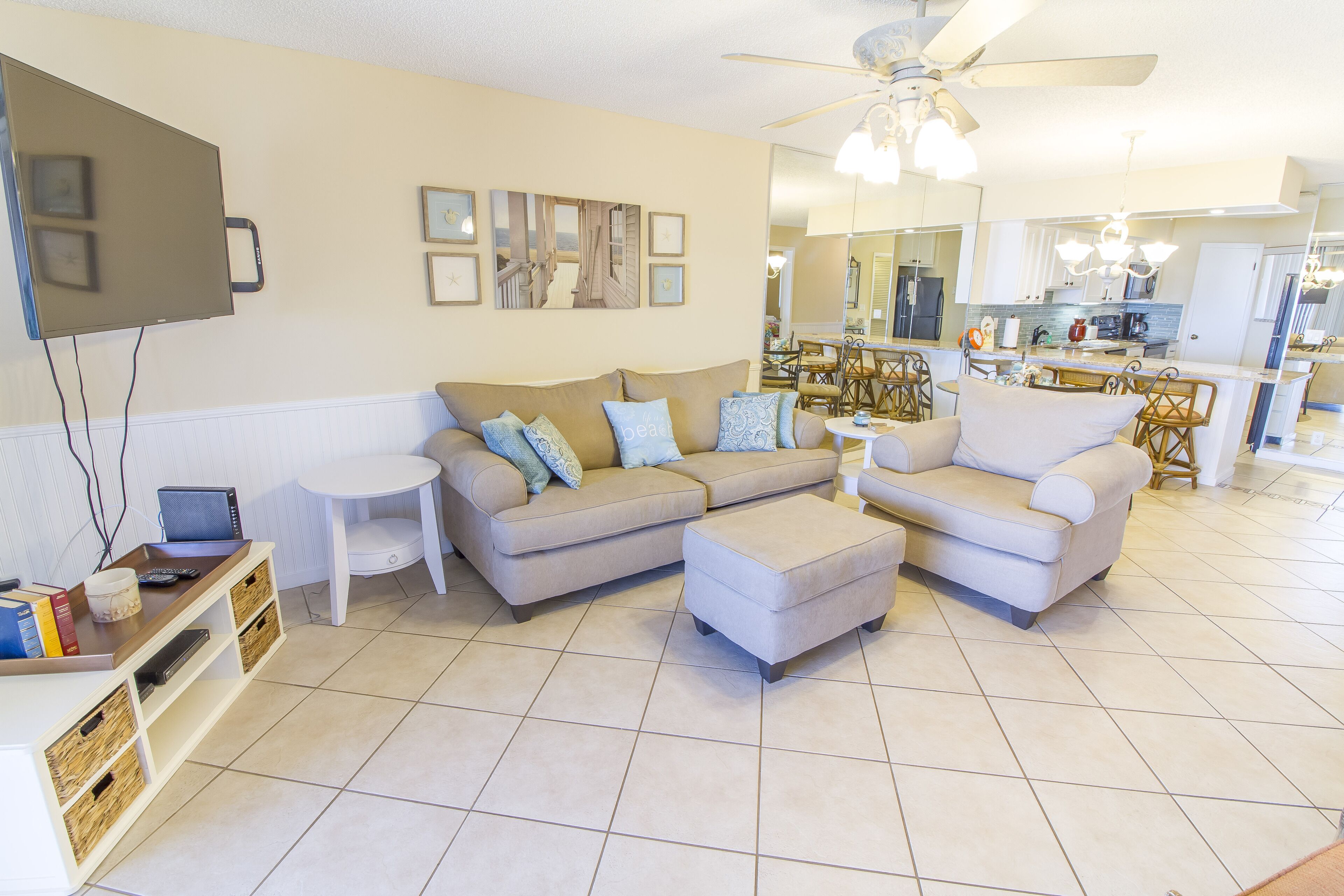 A living room with a beige sofa, a white armchair, and a coffee table. There is a TV mounted on the wall, a small side table, and a bookshelf.
