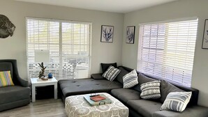 A modern living room with a grey sectional sofa, a patterned ottoman, and a small white side table with a lamp.