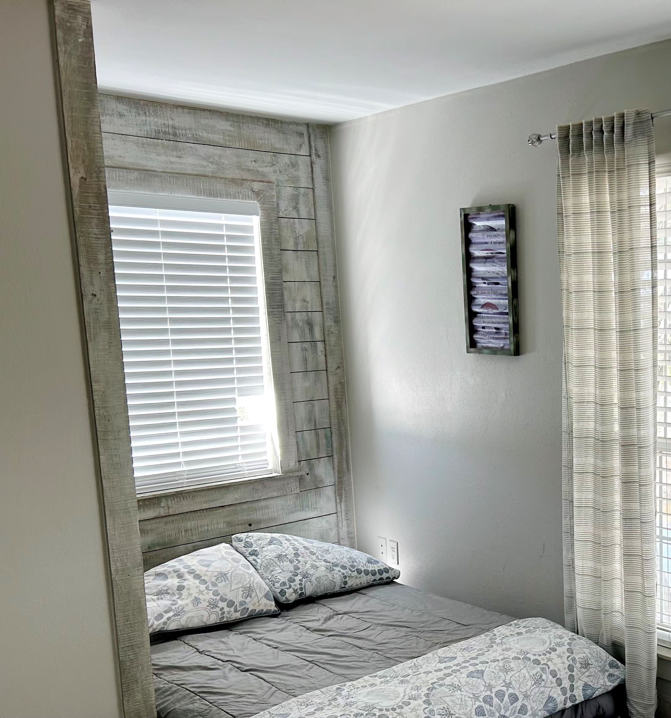 A bedroom with a window, a bed with patterned bedding, a wall-mounted light, and a curtain.