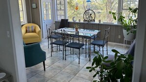 A dining area with a table and chairs, a yellow armchair, and a view of trees through large windows.