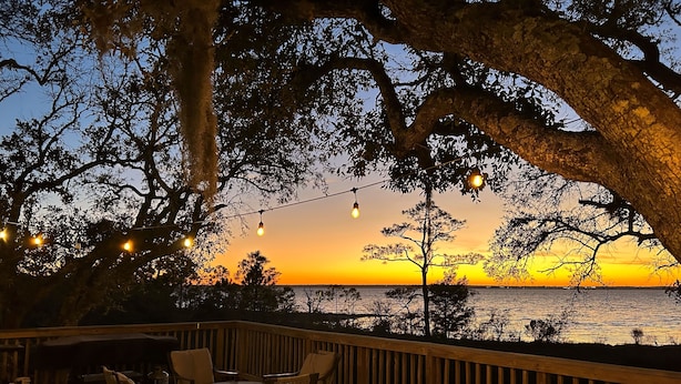 A wooden deck with outdoor furniture and string lights, overlooking a body of water at sunset.
