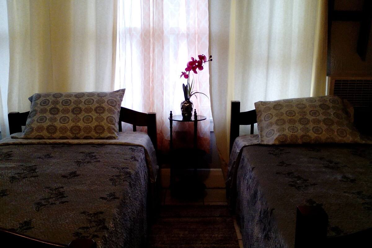A room with two beds, each with patterned bedding and matching pillows. There is a small table with a vase of flowers between the beds.