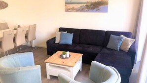 Un salon avec un canapé foncé, des fauteuils clairs, une petite table basse en bois et un tableau représentant une scène de plage.