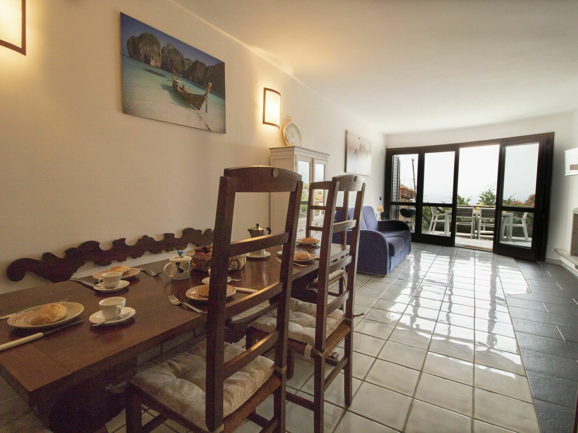 A dining area with a wooden table set for two, a painting of a coastal scene, and a view through large glass doors to an outdoor area with chairs.