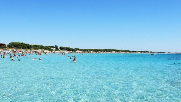 A beach with clear turquoise water, people swimming and sunbathing, and a distant shoreline with buildings. - Cosy apartment in villa for 4 people with A/C, WIFI, pool and terrace (Porto Cesareo)