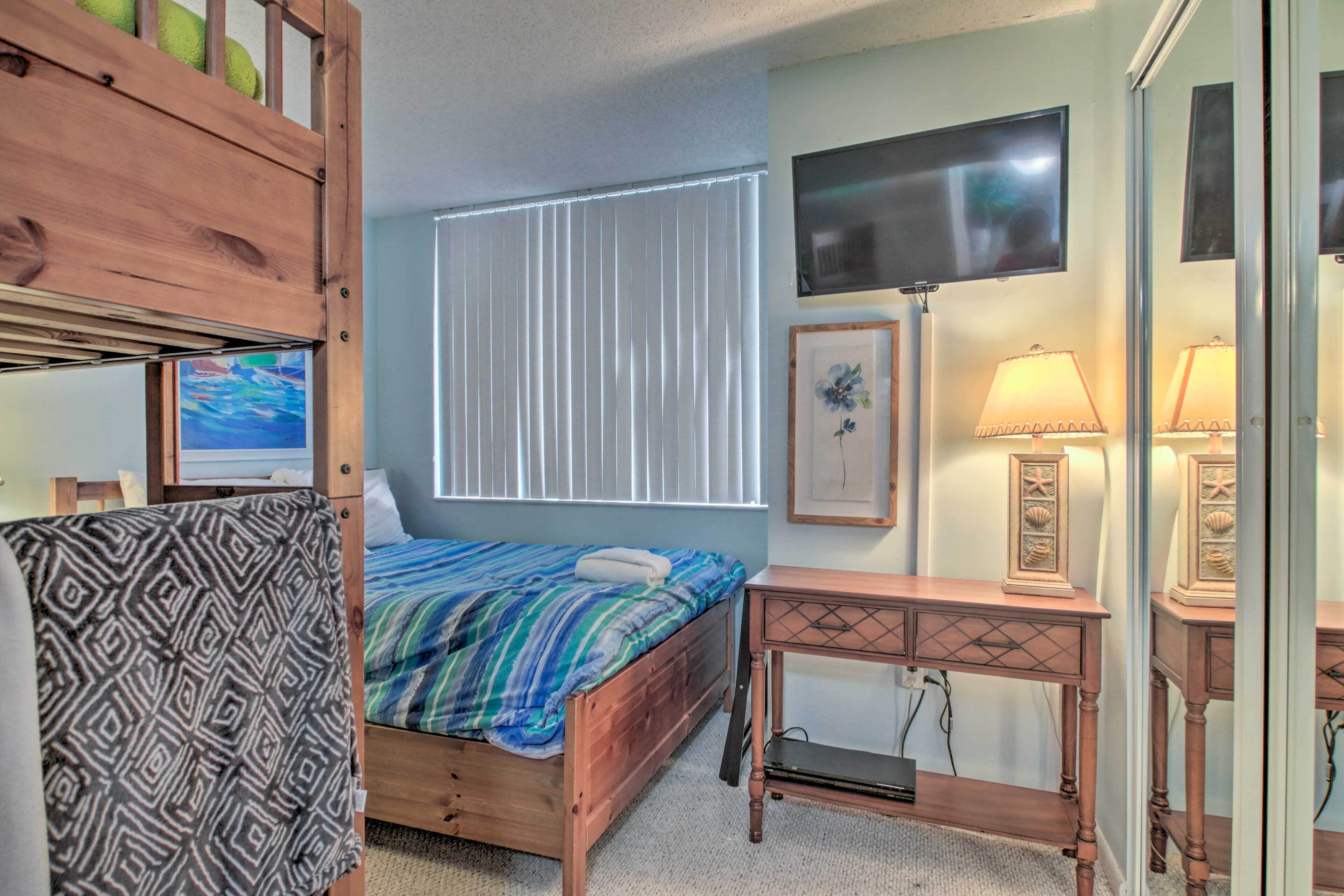 A bedroom with a bunk bed, a single bed with a striped blanket, a bedside table with a lamp, a TV mounted on the wall, and a window with blinds.
