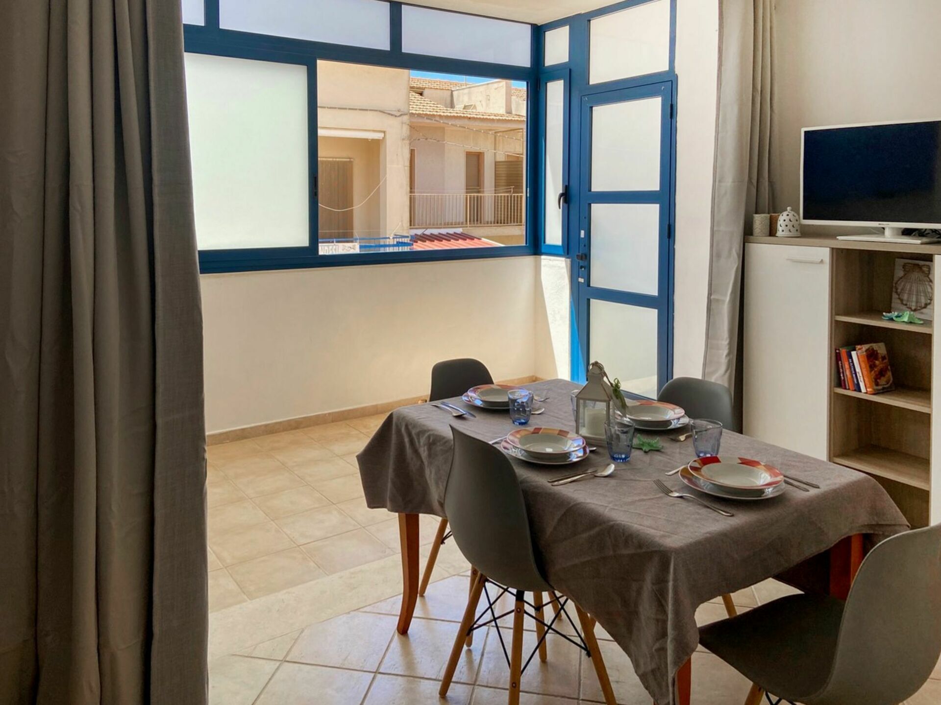 A dining area with a table set for four, a TV on a stand, and a bookshelf with books.