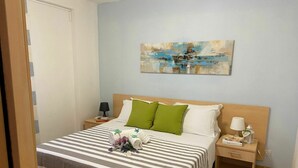 Ein ordentlich bezogenes Bett mit gestreifter Bettwäsche, ein grünes Kissen und eine Blumenkomposition. Darüber an der Wand hängt ein Gemälde, ein Nachttisch mit einer Lampe und eine Holzkommode neben dem Bett.