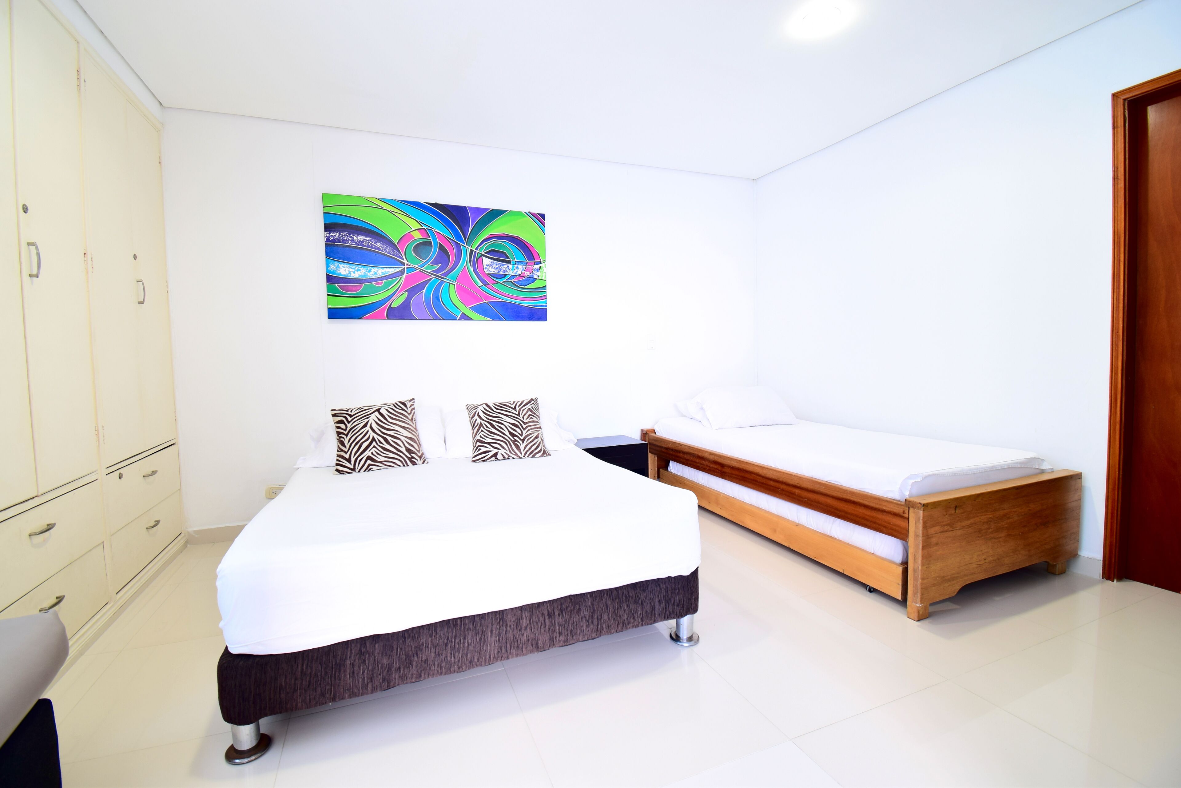 A bedroom with a large bed, a smaller bed, a wardrobe, and a colorful abstract painting on the wall.