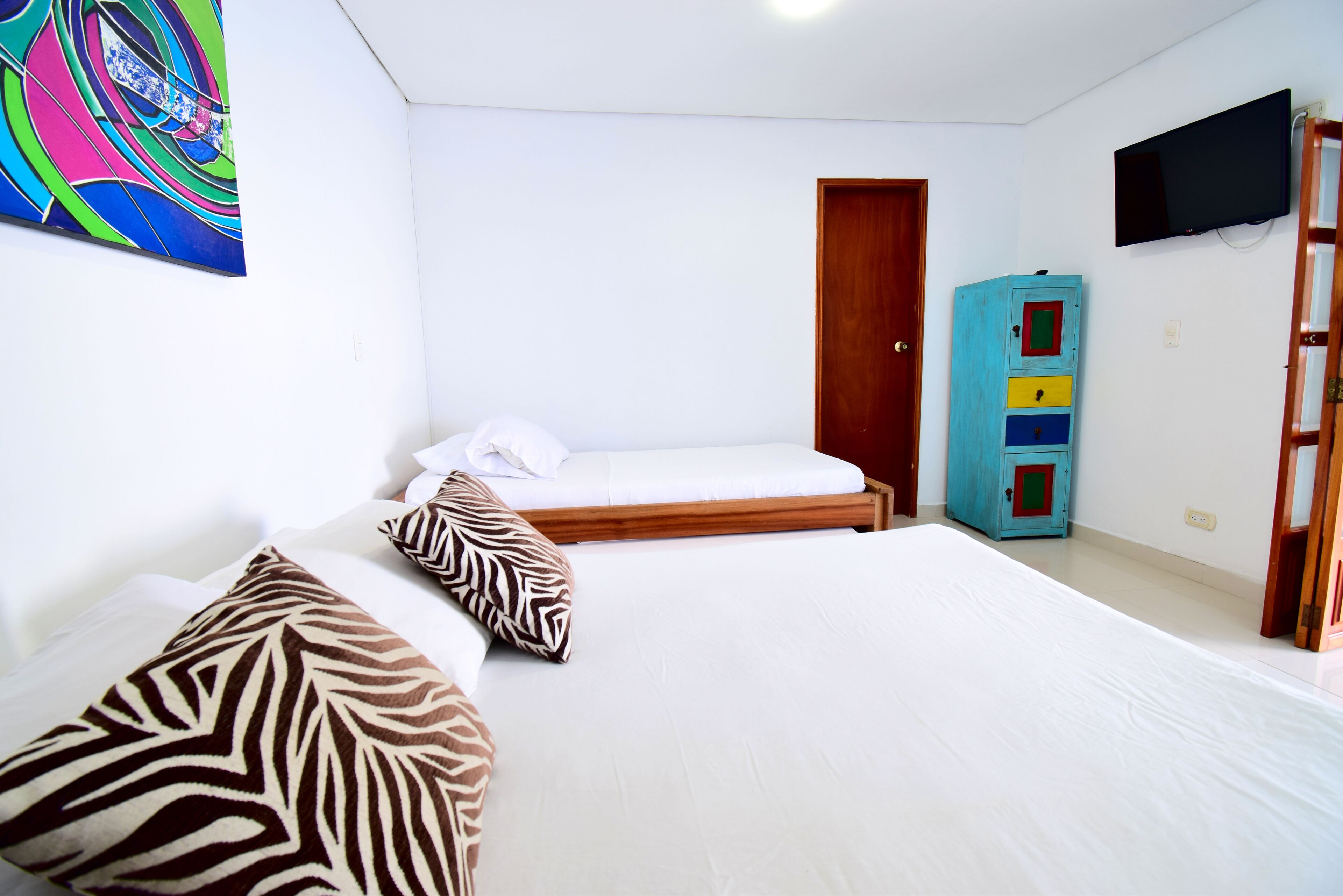 Habitación de hotel con dos camas, un cuadro abstracto colorido, un televisor y un mueble azul con cajones.