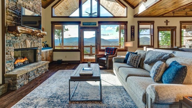 A spacious living room with a stone fireplace, wooden ceiling beams, and large windows offering mountain views. - Luxury 3BR Mountain Cabin • Hot Tub • Game Room • Outdoor Fireplace (Epworth)