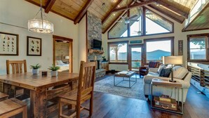 A spacious living area with a high ceiling, wooden beams, a stone fireplace, and large windows offering a view of the outdoors. - Luxury 3BR Mountain Cabin • Hot Tub • Game Room • Outdoor Fireplace (Epworth)