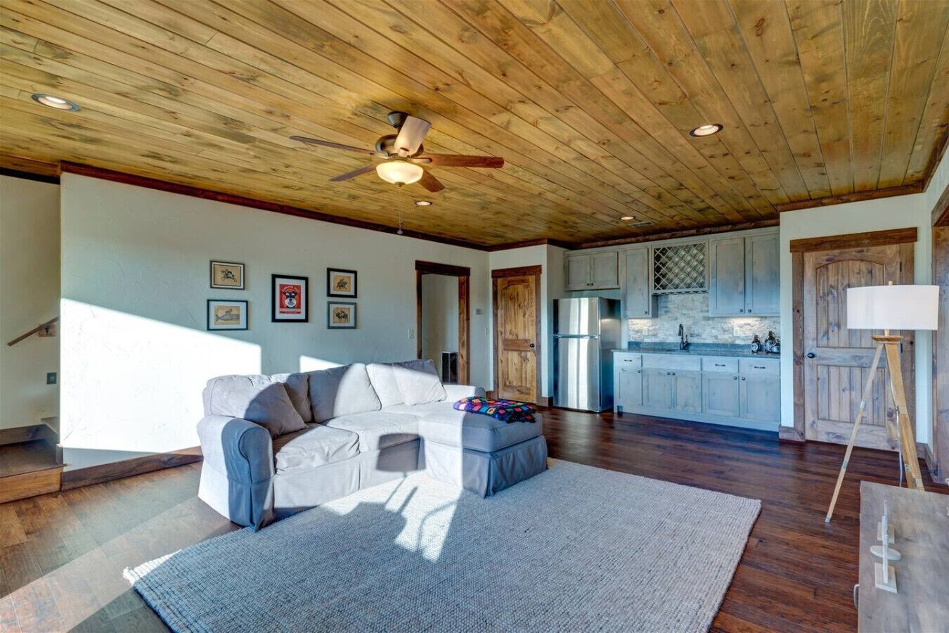 A spacious living room with a wooden ceiling, a ceiling fan, a grey sofa, a coffee table, and a kitchen area with wooden cabinets and a refrigerator.