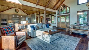 A spacious living area with a high ceiling, wooden beams, a large sofa, a coffee table, and a view of the kitchen and dining area. - Luxury 3BR Mountain Cabin • Hot Tub • Game Room • Outdoor Fireplace (Epworth)