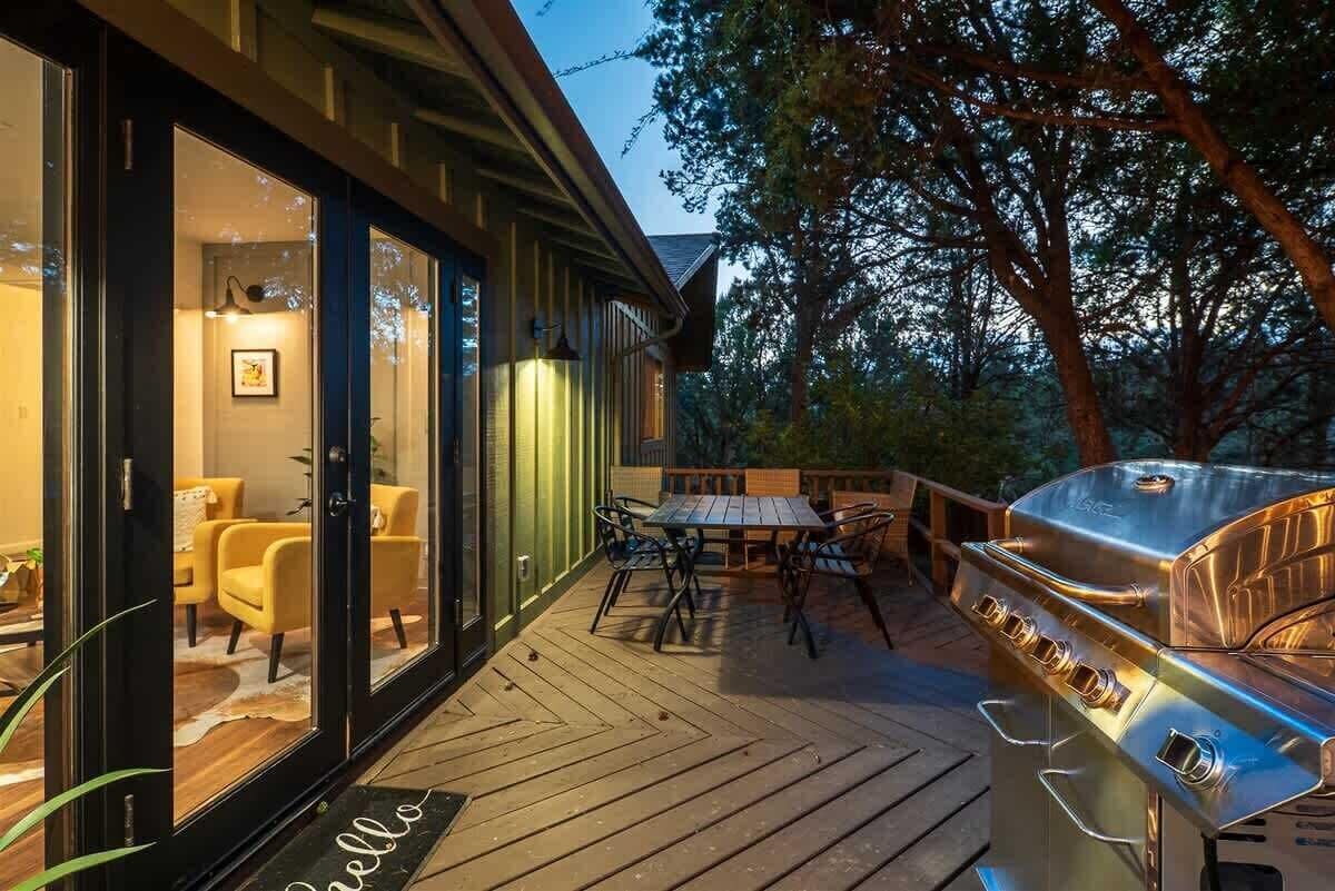 An outdoor deck with a grill, a glass door leading to a room with yellow chairs, and a wooden deck.