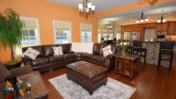 A spacious living room with a leather sectional sofa, a coffee table, and a rug. There is a dining area with a table and chairs, and a kitchen with wooden cabinets and a countertop.