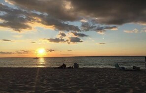 Ein Strand bei Sonnenuntergang, die Sonne tief am Horizont, ein Boot im Wasser und einige Personen sowie Stühle im Sand.