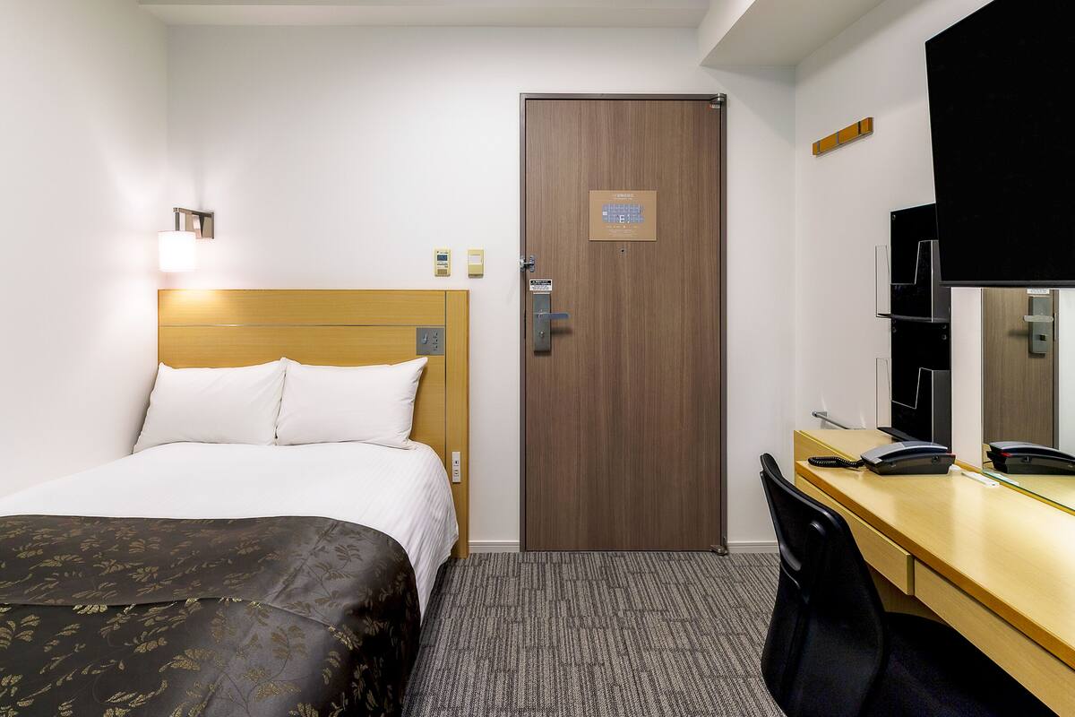 A hotel room with a bed, a desk, a chair, a door, and a mirror.
