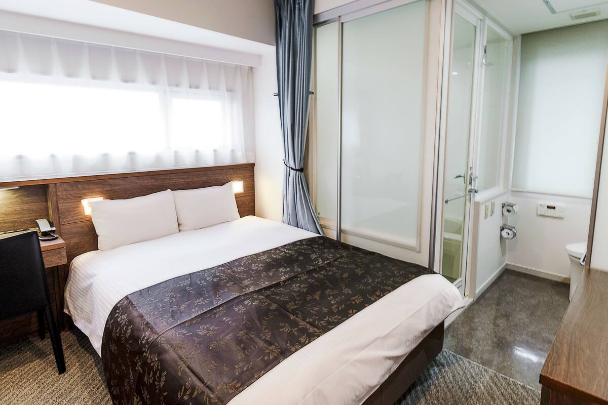 A hotel room with a bed, a desk, a telephone, a window with curtains, and a sliding door leading to a bathroom.