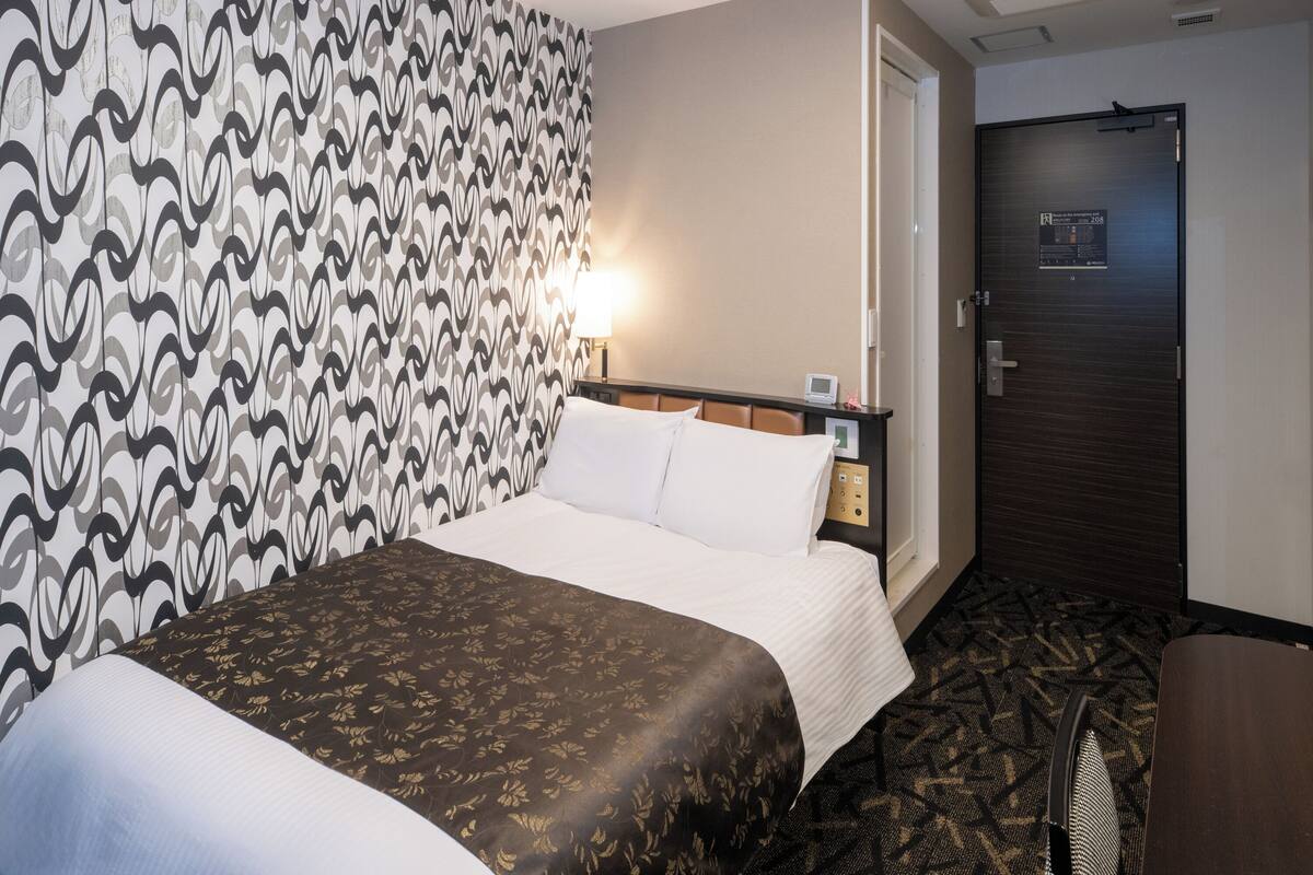 A hotel room with a patterned wallpaper, a bed with a brown and white bedspread, a bedside table with a lamp, and a door with a sign.