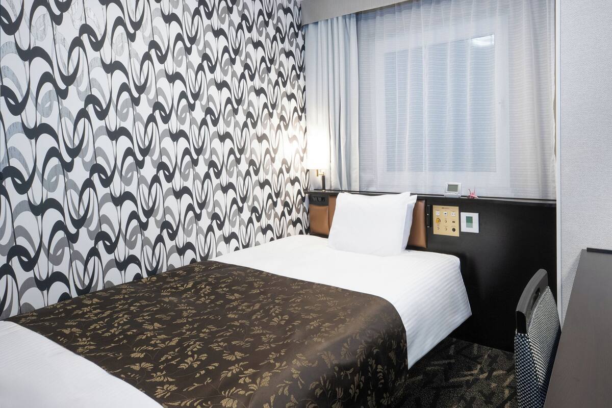 A hotel room with a patterned wall, a bed with a floral bedspread, a bedside table with a lamp, and a window with curtains.