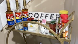 Carrito metálico dorado de tres niveles con botellas de sirope Torani, un letrero de café y varios utensilios de cocina.