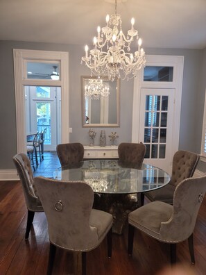 A dining room with a round glass table, four upholstered chairs, a chandelier, and a large mirror.