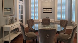 A dining room with a glass table surrounded by four upholstered chairs, a white console table with decorative items, a chandelier, and two windows with shutters. - Historic Hideaway with pool!  Close to beaches and downtown. (Galveston)