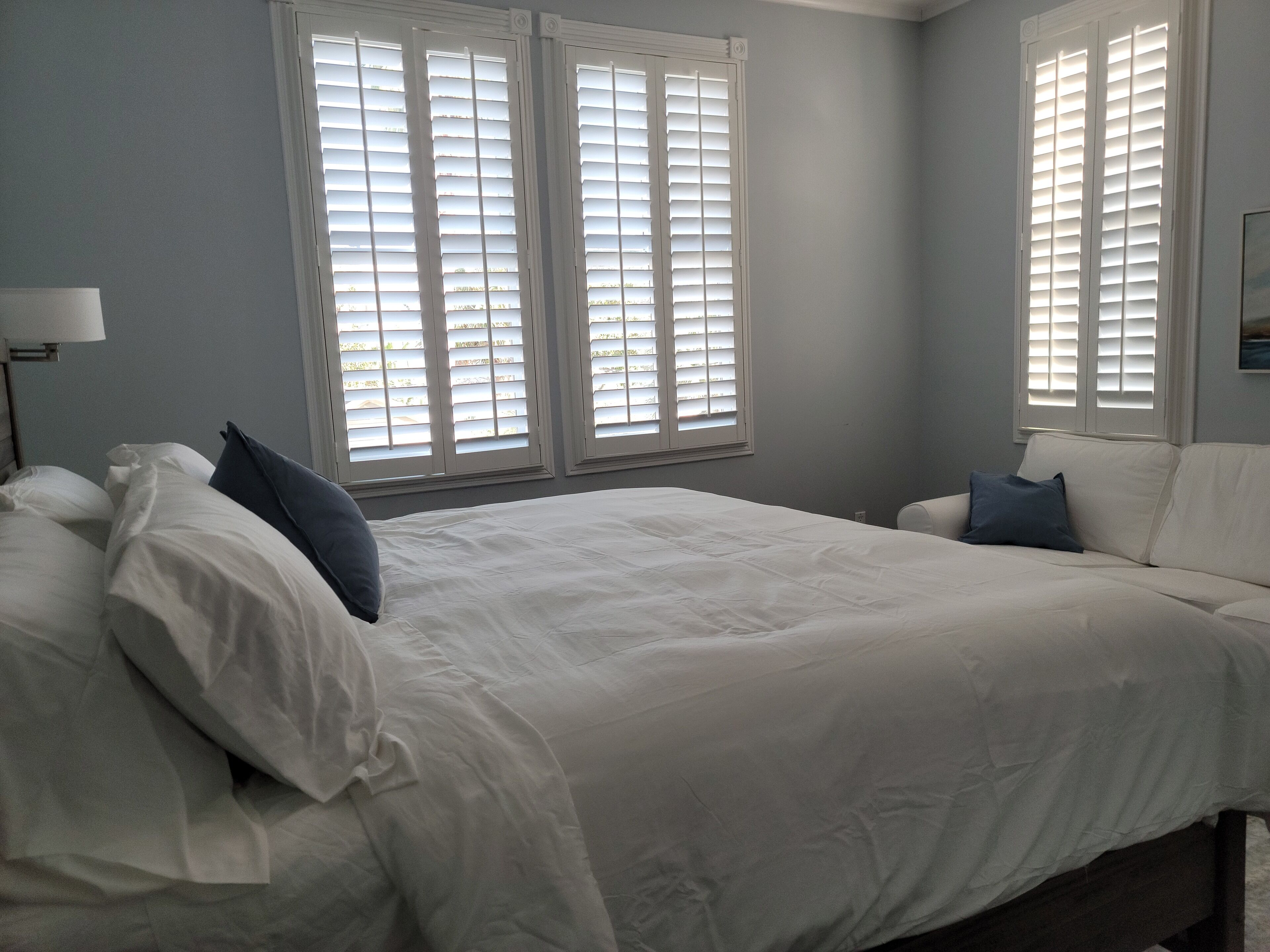 Un dormitorio con una cama grande, dos ventanas con persianas, una mesita de noche y una lámpara.