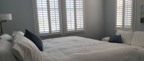 Un dormitorio con una cama grande, dos ventanas con persianas, una mesita de noche y una lámpara.