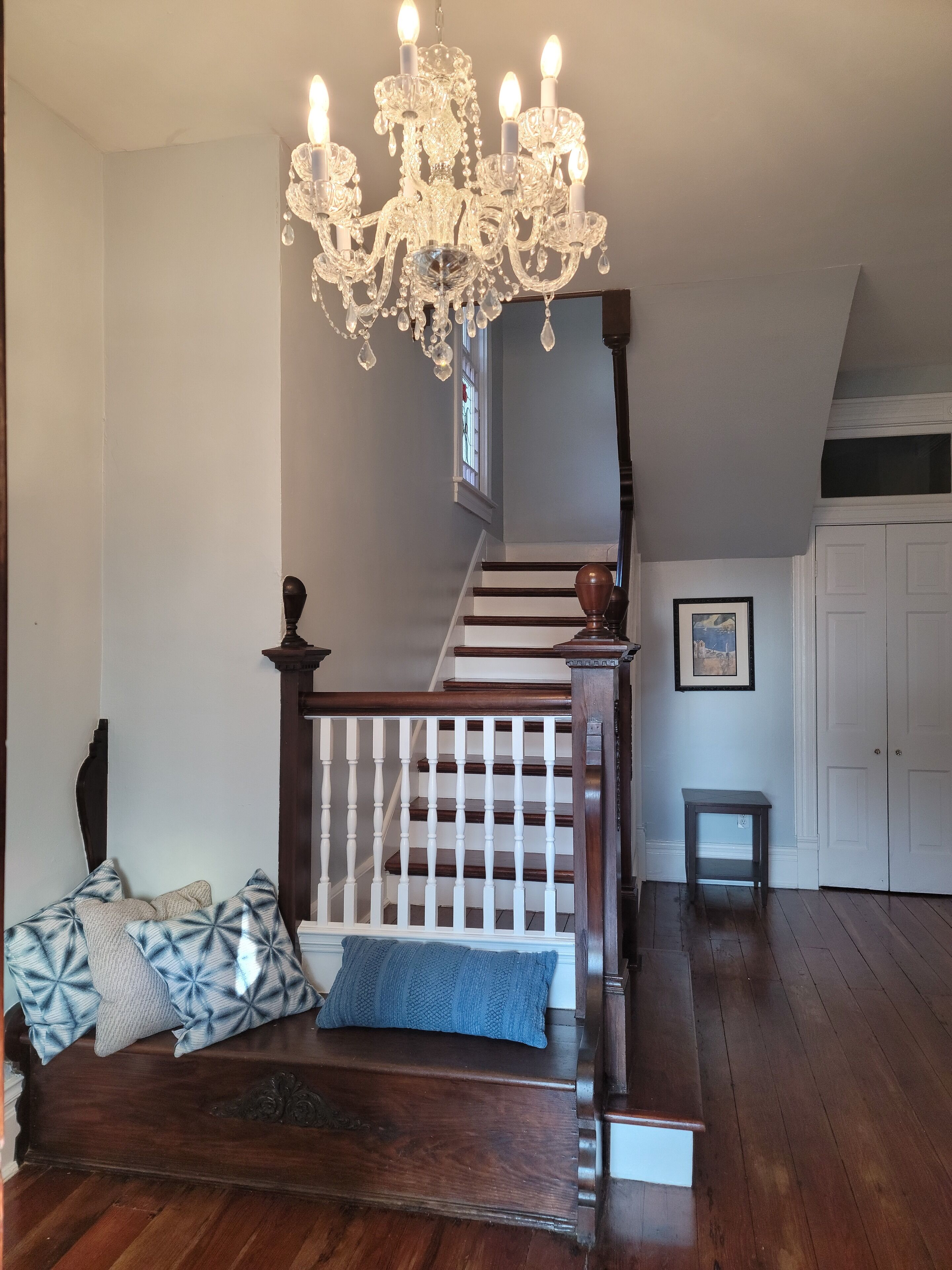 Un escalier avec une main courante en bois et des balustrades blanches, menant à un palier agrémenté d’un lustre et d’un tableau encadré au mur.