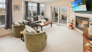A living room with a sofa, two green armchairs, a wooden coffee table, a fireplace, and a TV displaying a beach scene. - Condo close to hiking, river, with mountain views (Thornton)