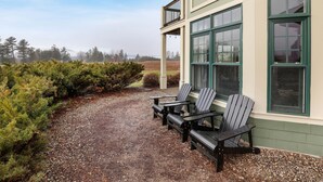 A covered outdoor seating area with Adirondack chairs and a table, adjacent to a building with large windows and a balcony. - Condo close to hiking, river, with mountain views (Thornton)