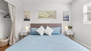 Quarto com cama, duas mesas de cabeceira, janela com persianas e quadro na parede.