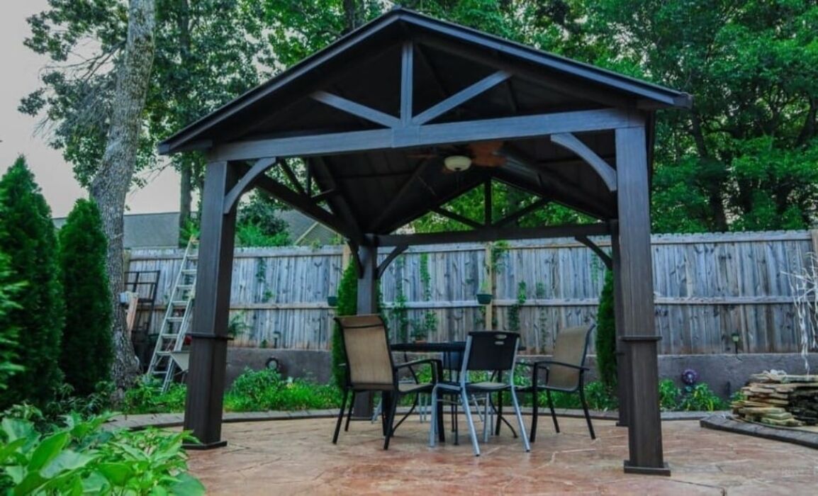 A covered patio area with a table and chairs, surrounded by a wooden fence and greenery.
