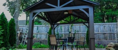 A covered patio area with a table and chairs, surrounded by a wooden fence and greenery.