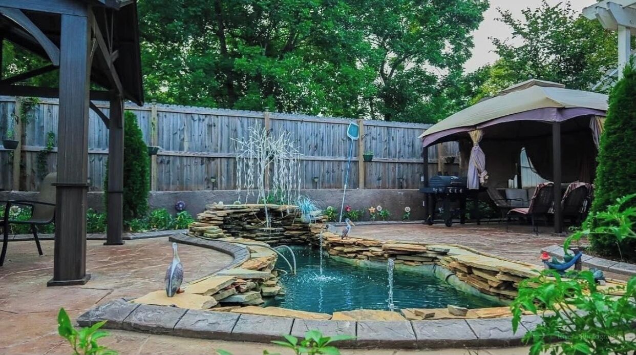 A backyard with a stone fountain, a wooden deck, and a covered seating area.