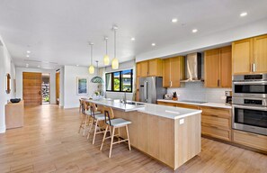Une cuisine moderne dotée d’un grand îlot central, de meubles en bois, d’appareils électroménagers en acier inoxydable et de luminaires suspendus.