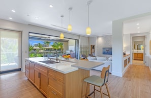 A modern kitchen with a large island, wooden cabinets, and a view of a balcony with palm trees.