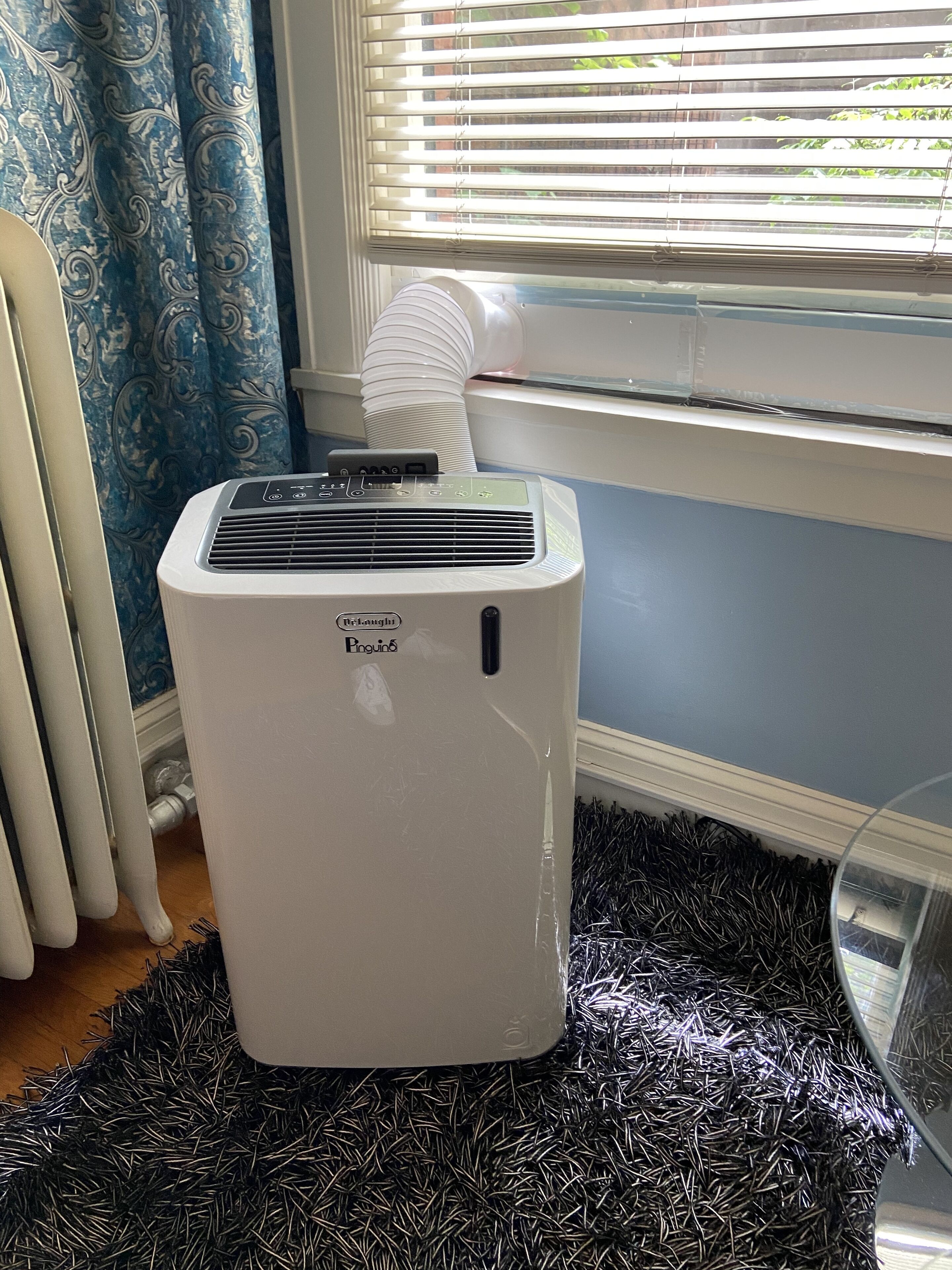 Un climatiseur portatif blanc se trouve dans une pièce dotée d’une fenêtre, d’un radiateur et d’un rideau à motifs.