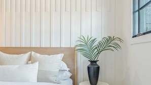 Une chambre à coucher avec une tête de lit en bois, un lit blanc, un tabouret rond blanc et une plante en pot.