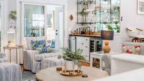 A modern living room with striped sofas, a round coffee table, and a kitchen area with glass shelving. - Goin' Coastal: East Beach 5BR Retreat | Private Pool | Steps to Beach | Sleeps 12 (Saint Simons Island)