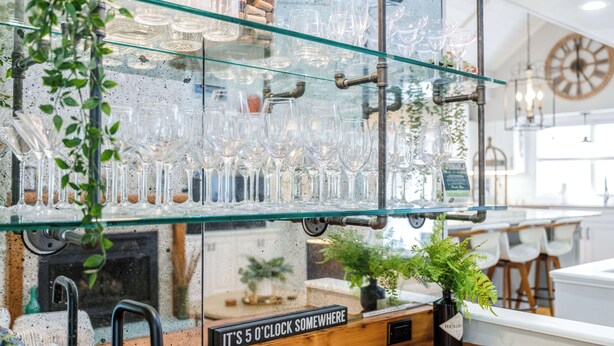 A bar area with glass shelves holding various glassware and bottles, a clock, and a sign that reads 'It's 5 o'clock somewhere.' - Goin' Coastal: East Beach 5BR Retreat | Private Pool | Steps to Beach | Sleeps 12 (Saint Simons Island)