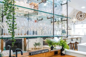 A bar area with glass shelves holding various glassware and bottles, a clock, and a sign that reads 'It's 5 o'clock somewhere.'